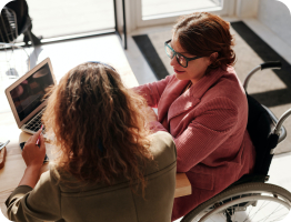 Two women at a desk looking at a laptop. One is using a wheelchair and wearing a pink jacket and glasses.