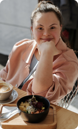 A young woman wearing a pink jacket and a ponytail is smiling at the camera with her hand resting under her chin