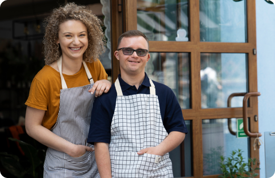A young woman with curly hair and a young man with brown hair and glasses standing together and both smiling and wearing aprons.