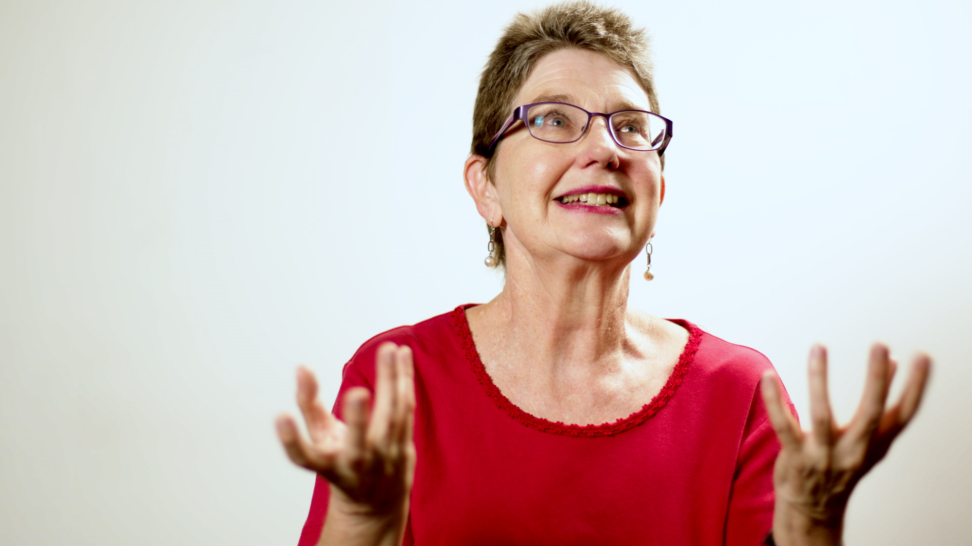 A lady wearing a red shirt and glasses with her hands open gesturing.