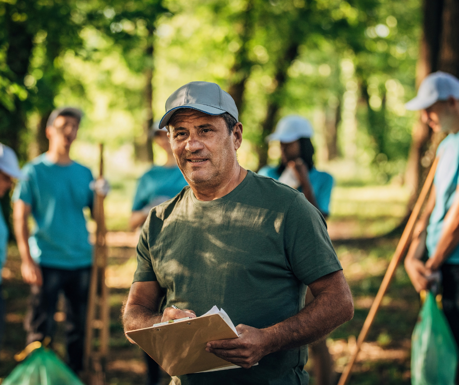 An older man in a park with a clipboard. There are 5 volunteers in the background gardening