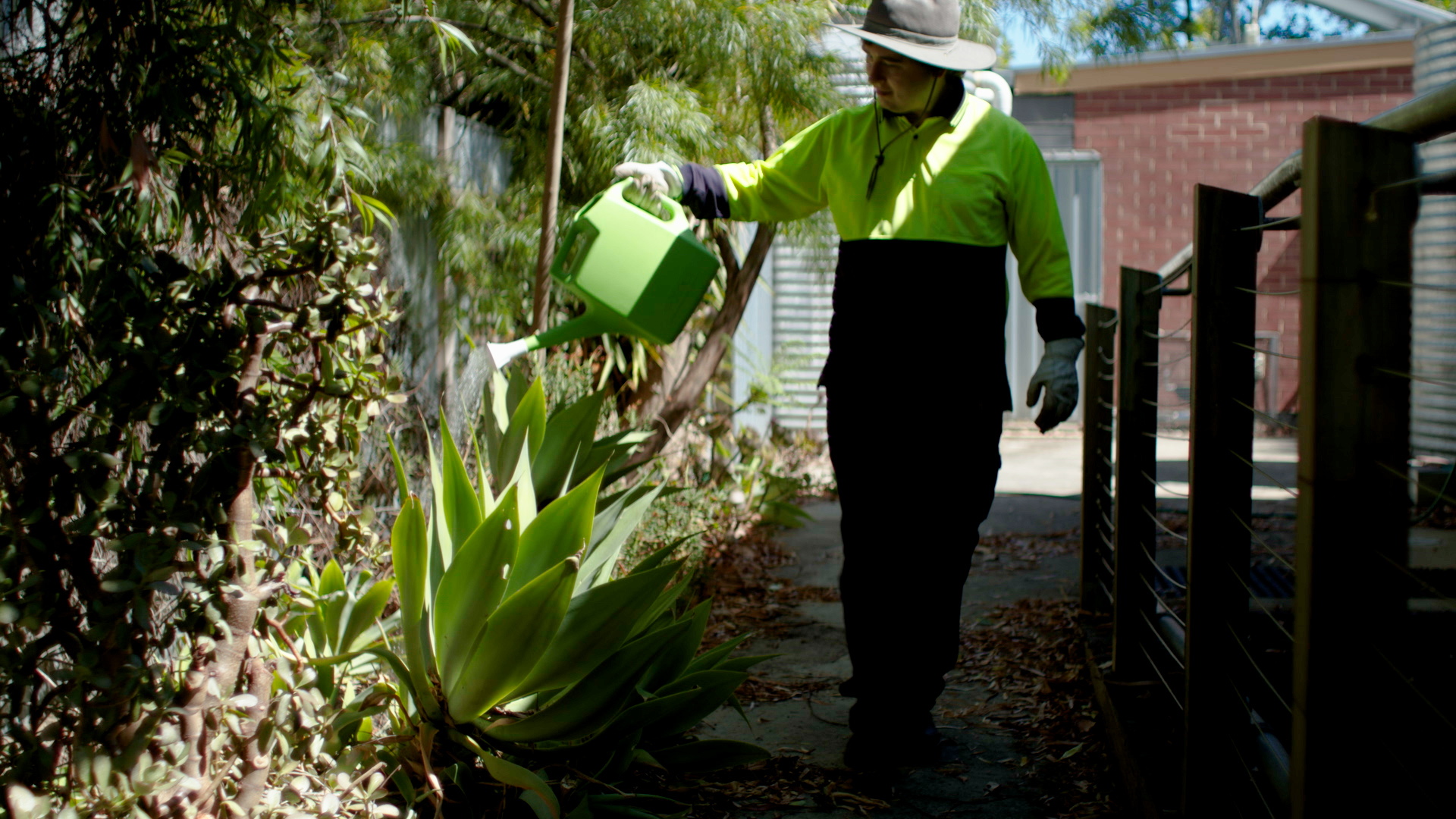 A young man wearing high viz long sleeved top and gardening gloves, watering plants outside with a watering can.