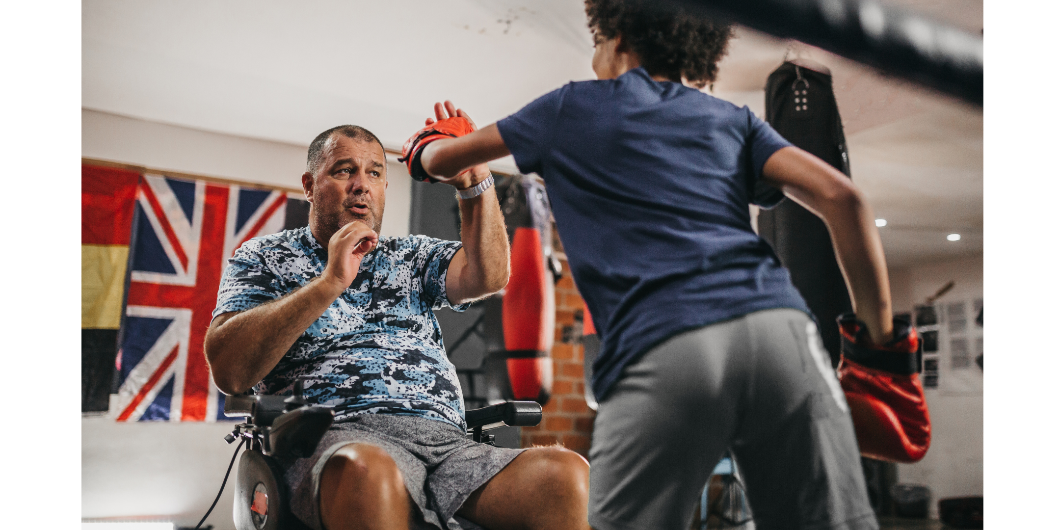 A young person is practicing boxing. An older man, using a wheelchair is helping them by holding his hands up in front of him