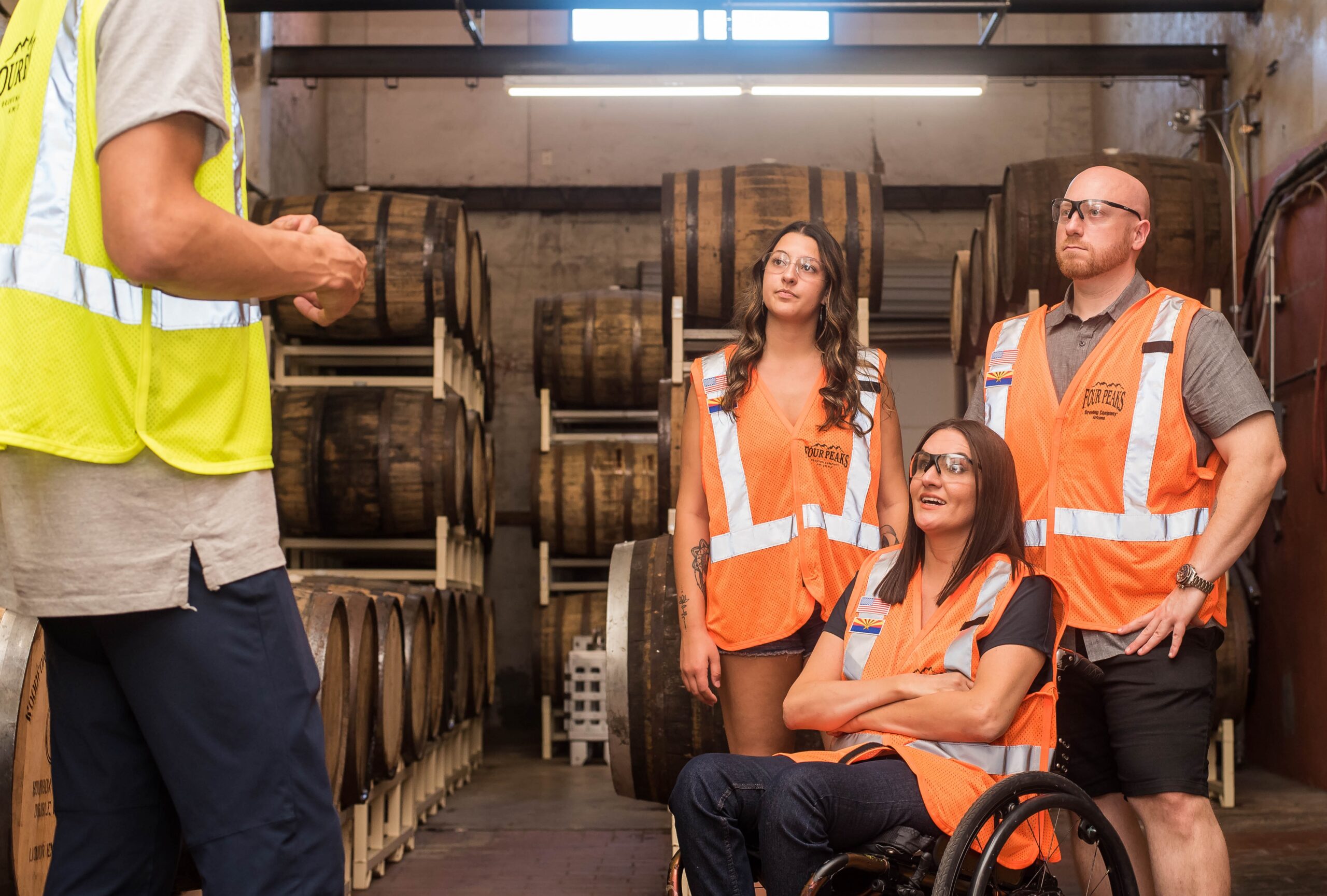 Two women and one man wearing orange high vis vests listening to another man give instructions. One is using a wheelchair .