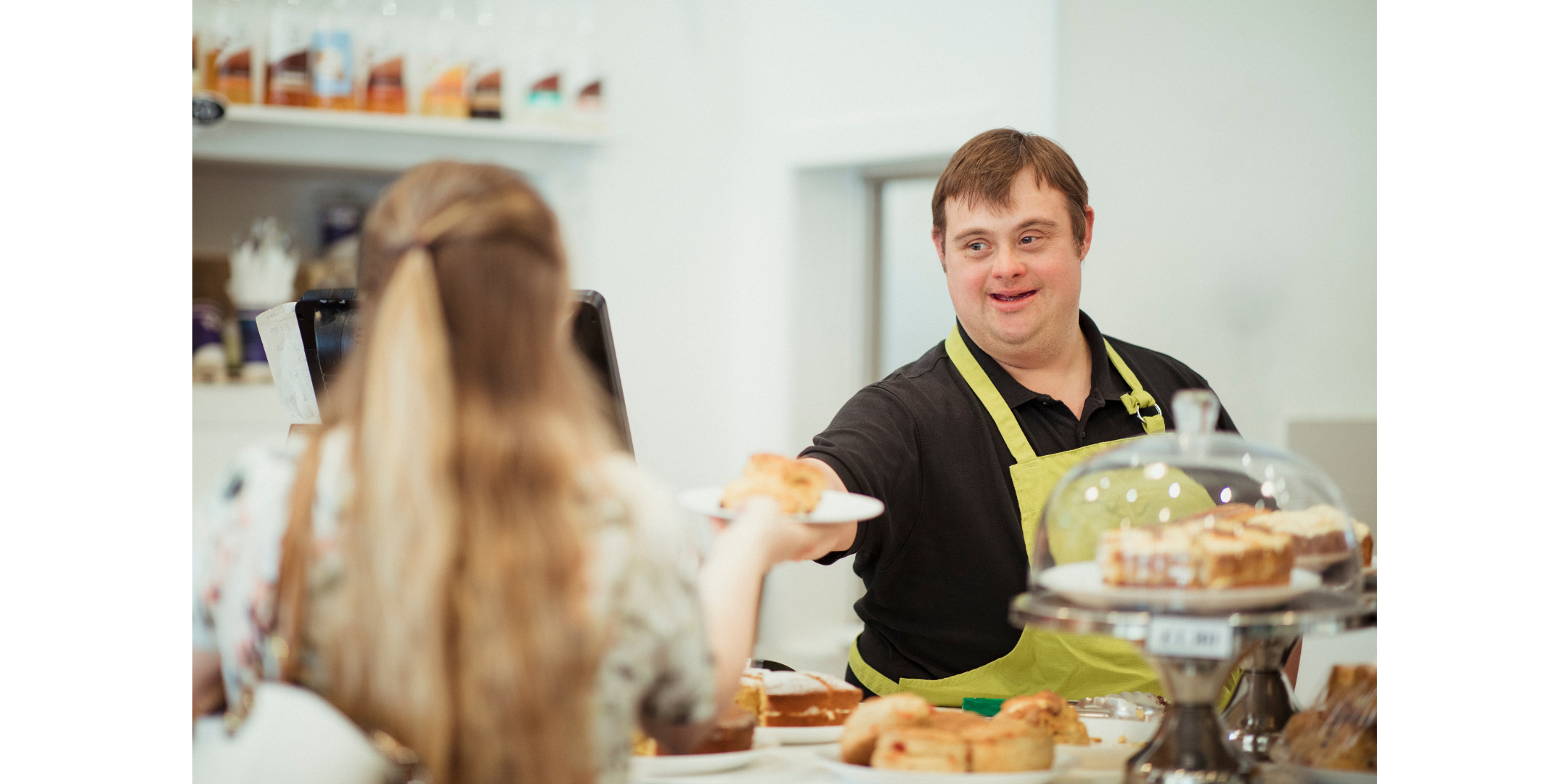 A young man with brown hair and wearing a yellow apron serving a scone to a lady across a counter