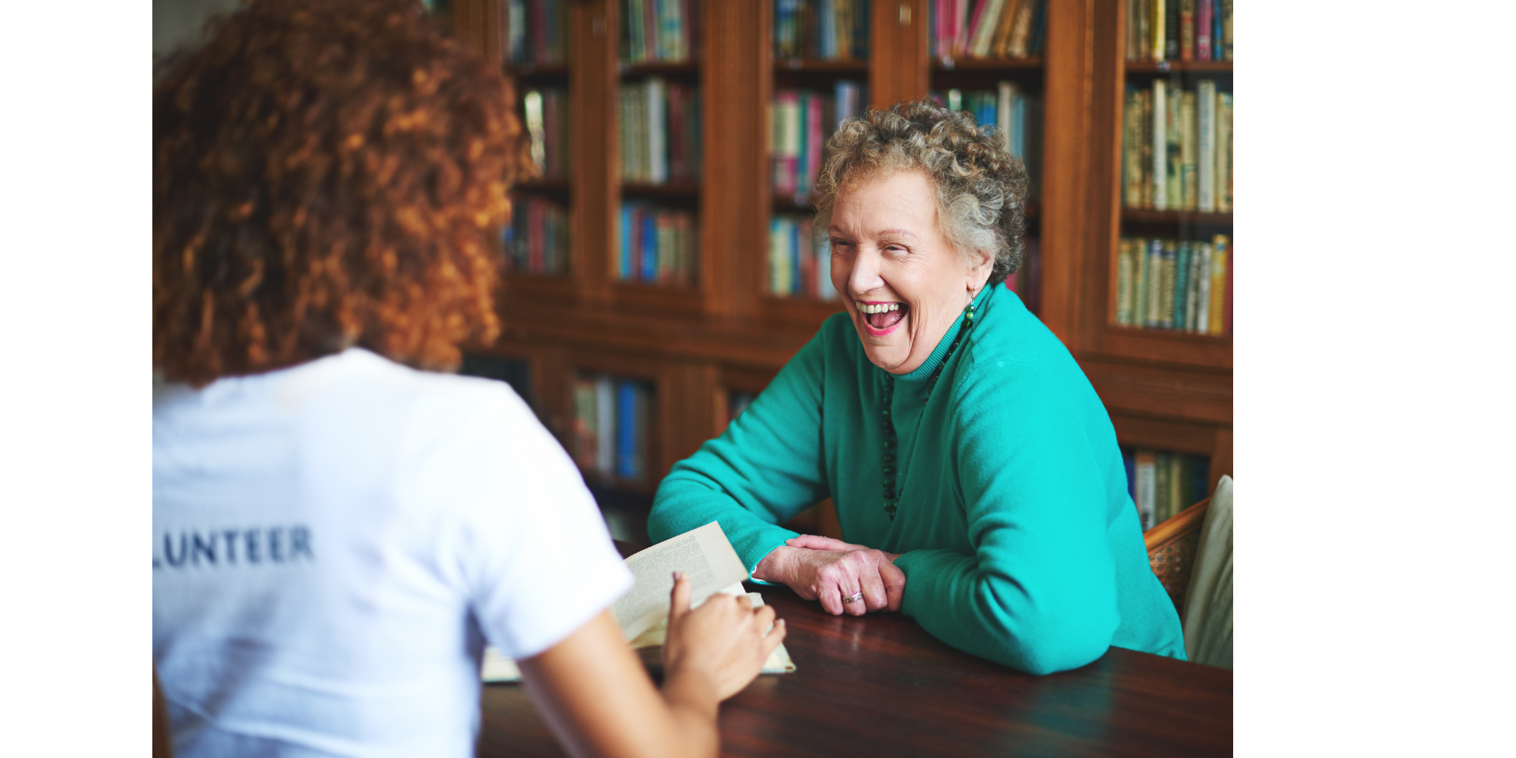 Laughing older woman sitting in a library opposite a woman wearing a T-shirt with volunteer written on the back