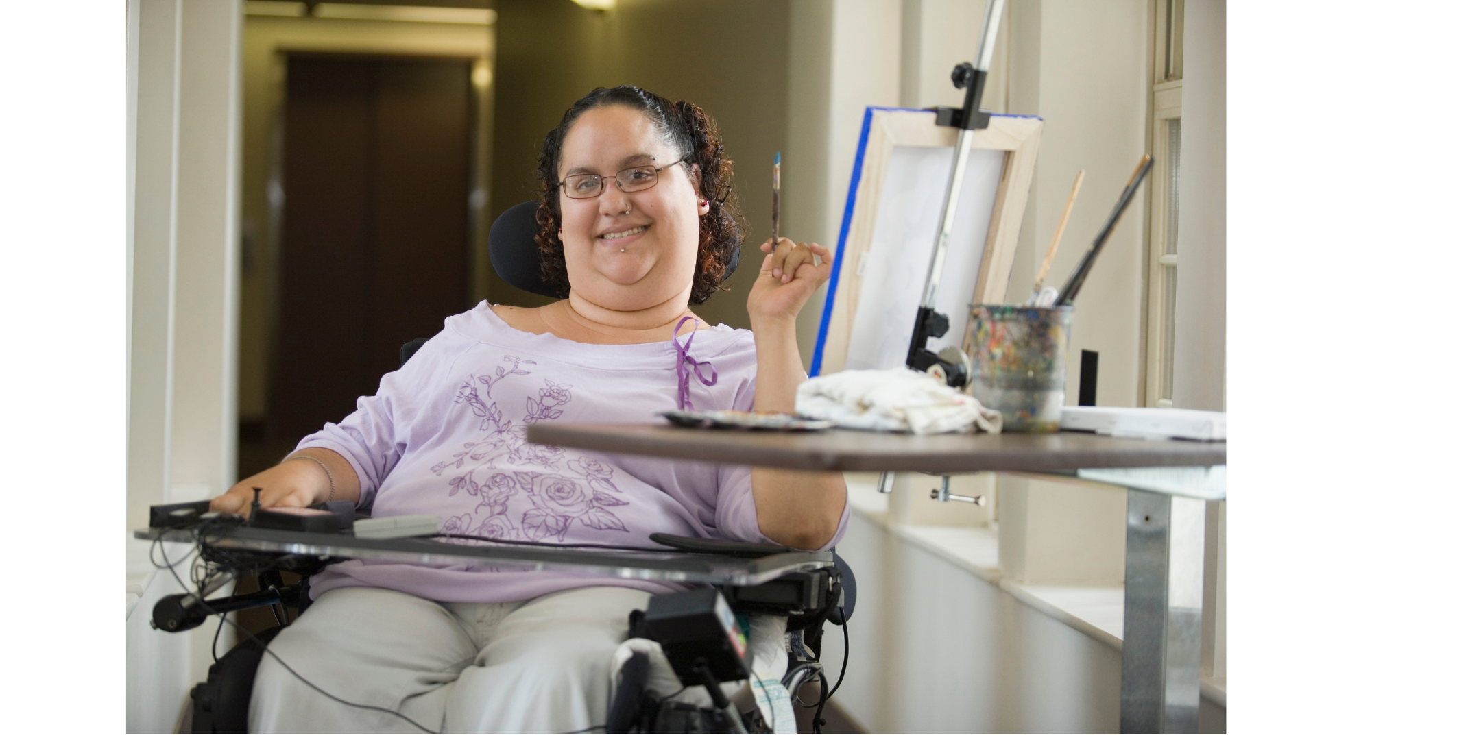 smiling woman with brown curly hair and glasses. sitting in her wheelchair besides an easel with a paintbrush in hand