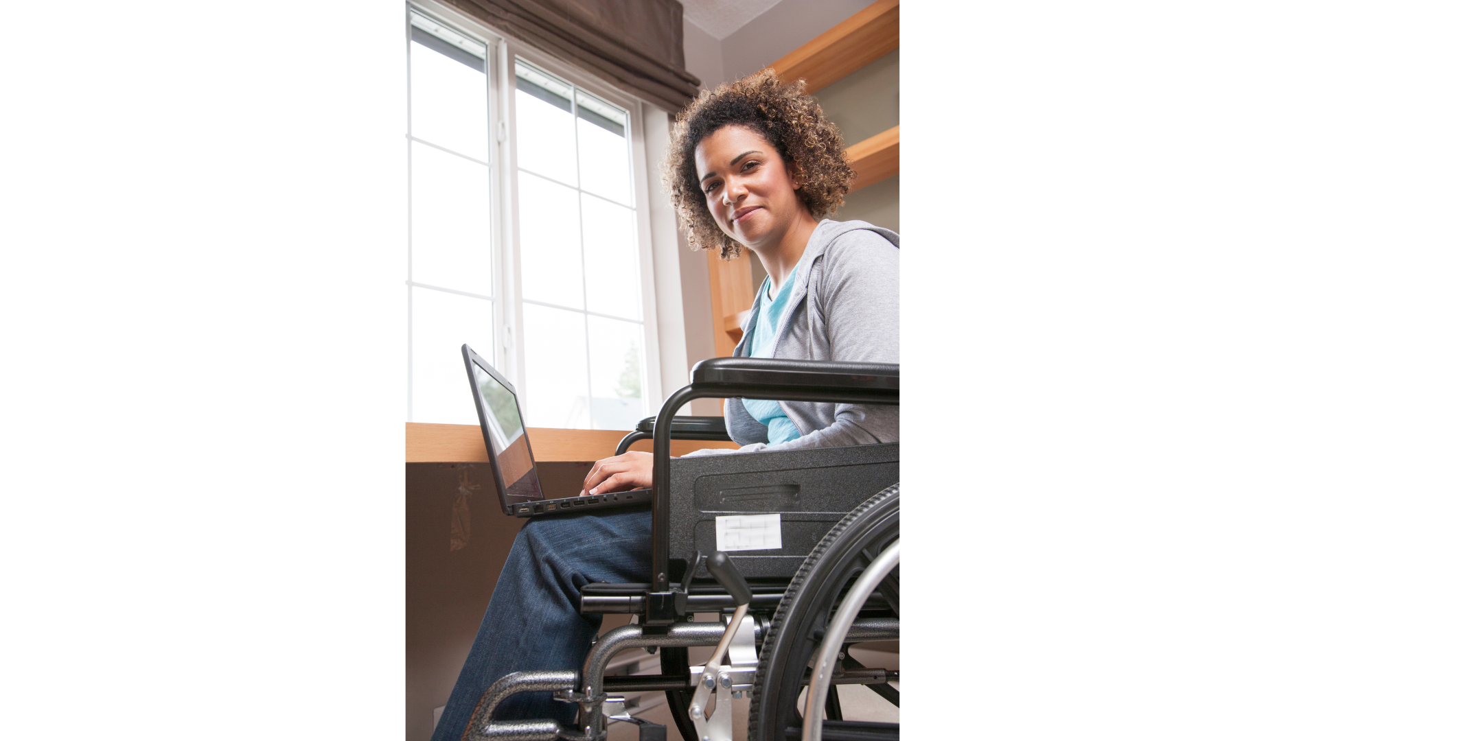 young woman with brown curly hair in wheelchair using laptop. wearing jeans and a grey sweatshirt jacket.